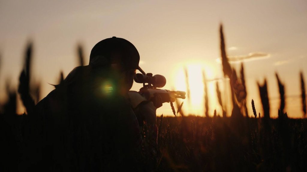 A person stands in a field holding a rifle surrounded by tall grass and open space