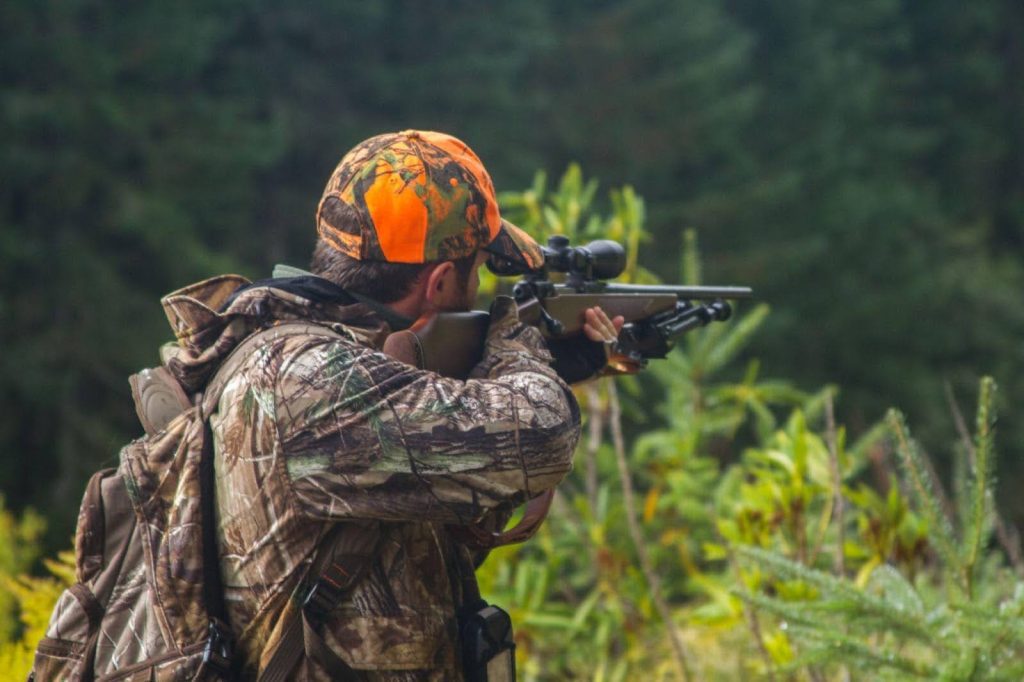 In a vast field, an individual is positioned with a rifle ready for action amidst the natural landscape