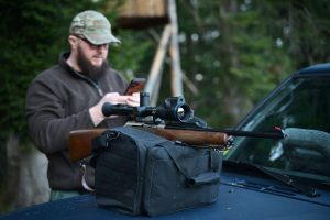 A man with a beard and a rifle checks his phone appearing focused in a natural setting