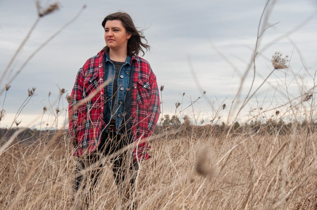 Woman wearing a red plaid jacket standing in a field of dry, tall grass under an overcast sky.