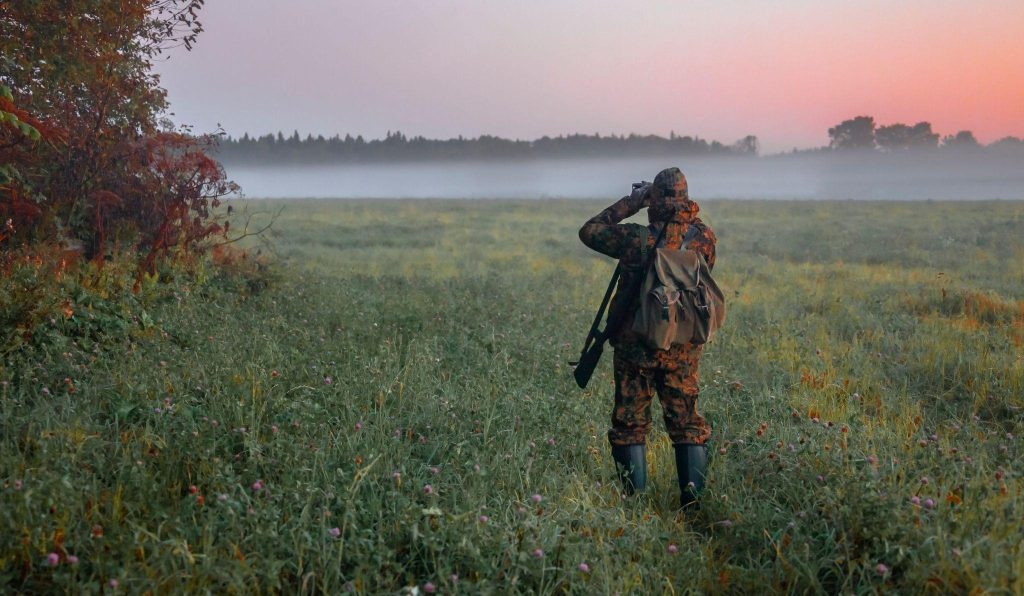 A man dressed in camouflage stands in a grassy field gripping a gun and looking intently ahead