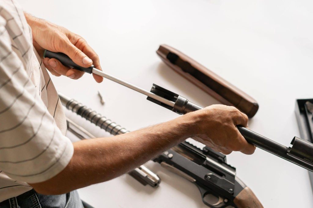 A person is cleaning a disassembled gun with a cleaning rod on a white surface