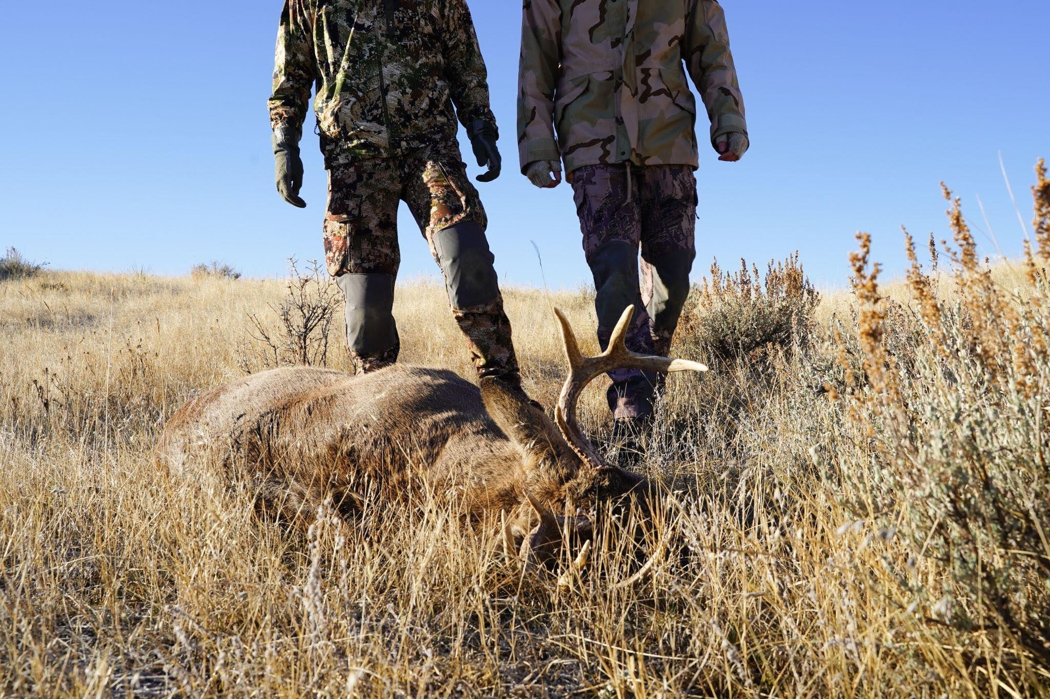 Two men pose next to a lifeless deer showcasing their successful hunt in a forested environment