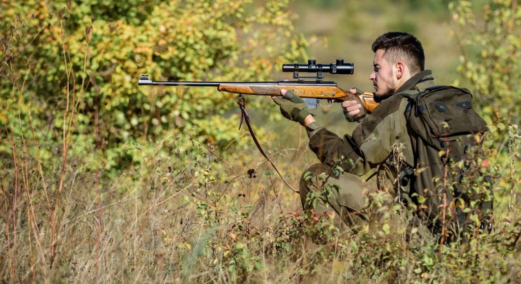 A man shooting a rifle in a field during a guided hunting trip, focusing on long-range hunting and choosing optics