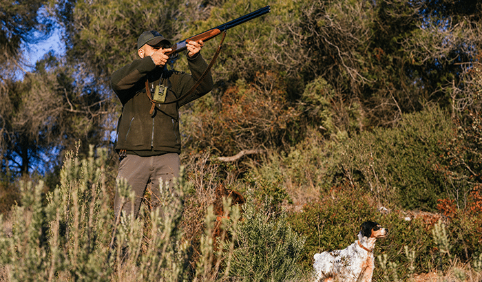 A hunter in camouflage gear holding a rifle, walking with a loyal dog in a forest.
