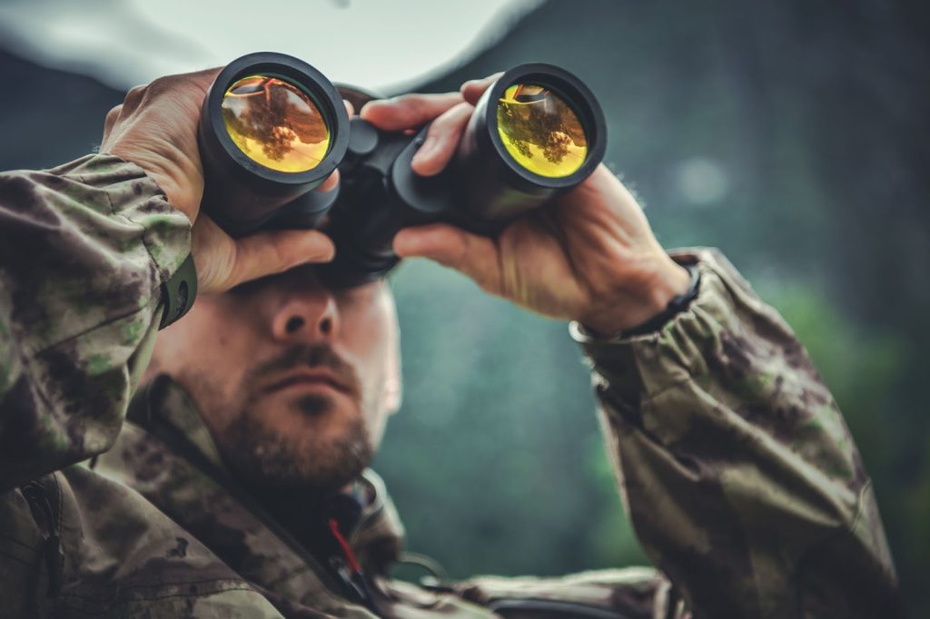 A person in camouflage using binoculars in a natural outdoor setting.