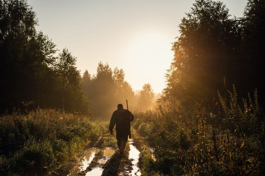 A person with a rifle walks down a muddy forest path at sunrise.