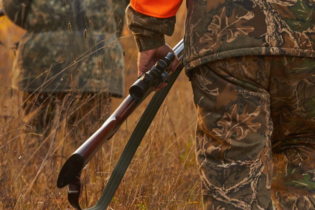 Hunter in camouflage holding a rifle in a grassy field.