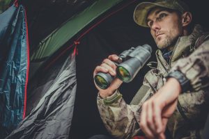 Man in camouflage holding binoculars inside a tent.
