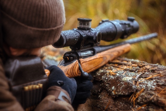 A hunter in winter gear aims a bolt-action rifle with a large scope, resting on a log in a forest setting.