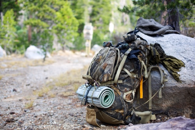 Camouflage backpack with sleeping pad, leaning against a boulder in a forest campsite.