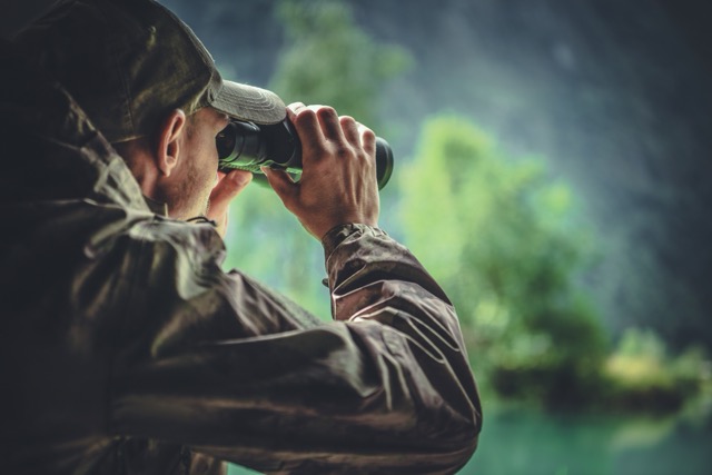 Man in camouflage jacket looking through binoculars in a forest.