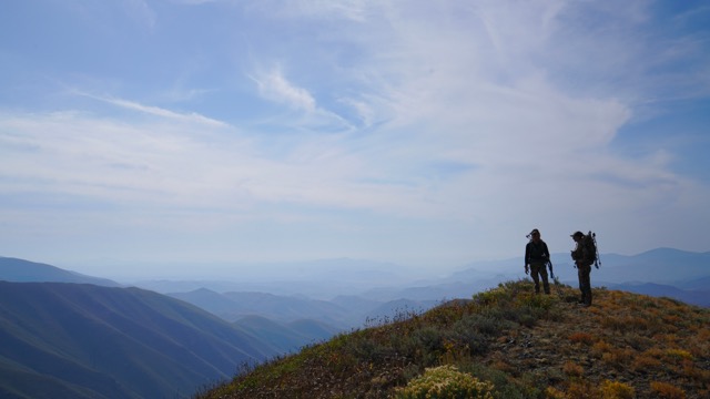 Two people stand atop a mountain, gazing over a vast valley below, surrounded by scenic landscapes.