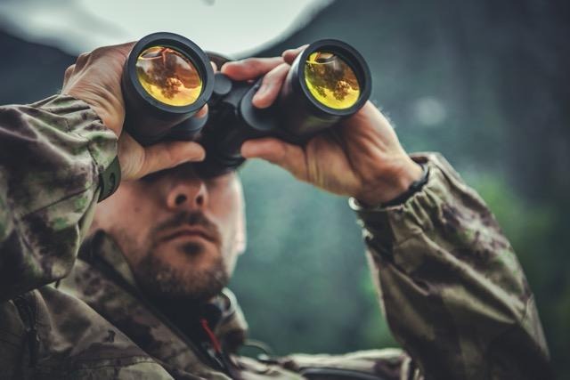 Close-up of a person in camouflage clothing looking through binoculars with a forest landscape reflected in the lenses.