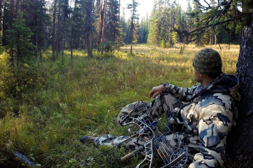 A man sits in a wooded area holding a bow surrounded by trees and natural foliage