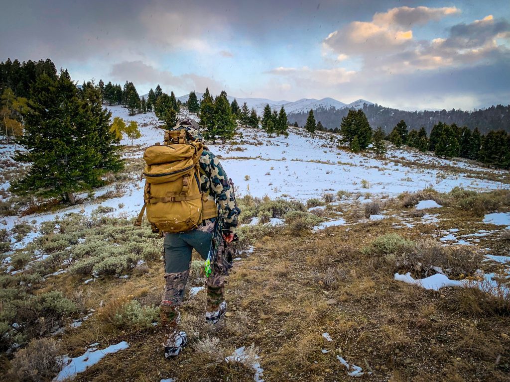 Rear view of a hunter wearing camouflage gear and a large tan backpack trekking through a snowy mountain landscape with evergreen trees.