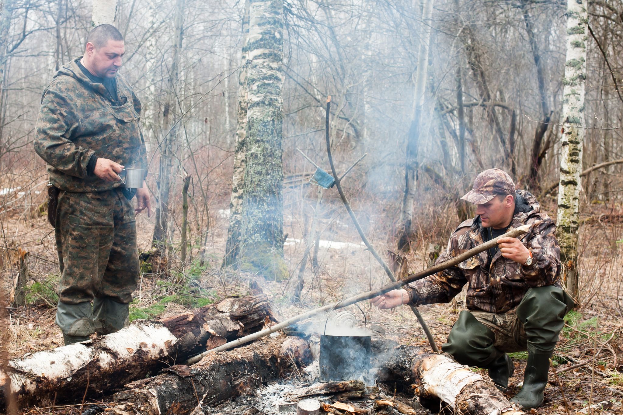 Two men in camouflage clothing cooking over a campfire in a wooded area.