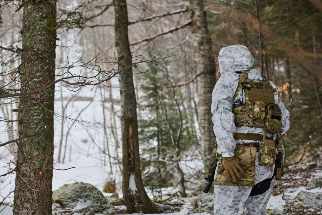 A man dressed in camouflage gear stands in a snowy landscape blending into the winter environment