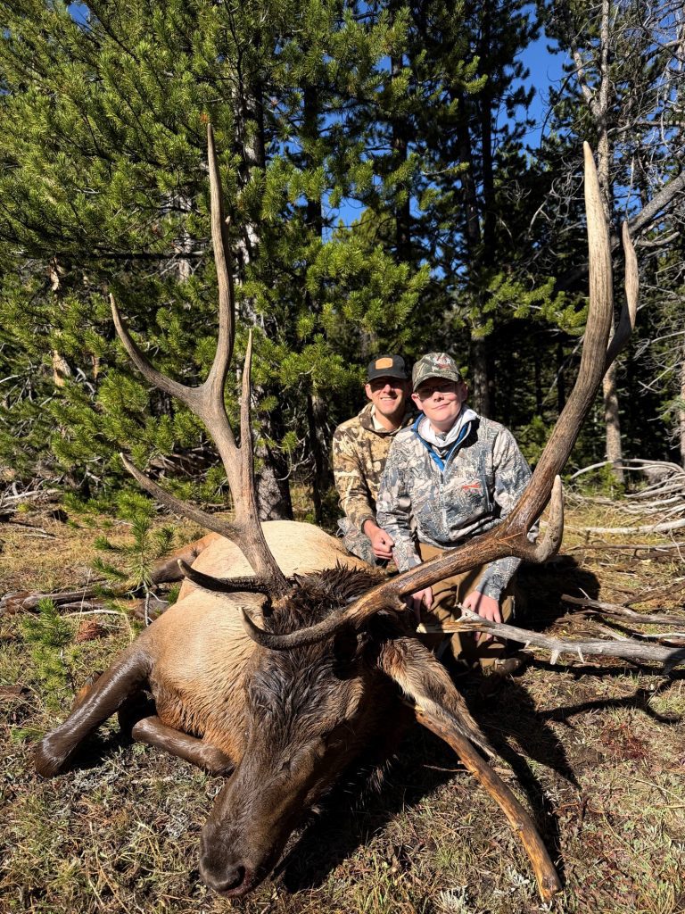 Two hunters in camouflage gear posing behind a harvested bull elk with large antlers in the woods.
