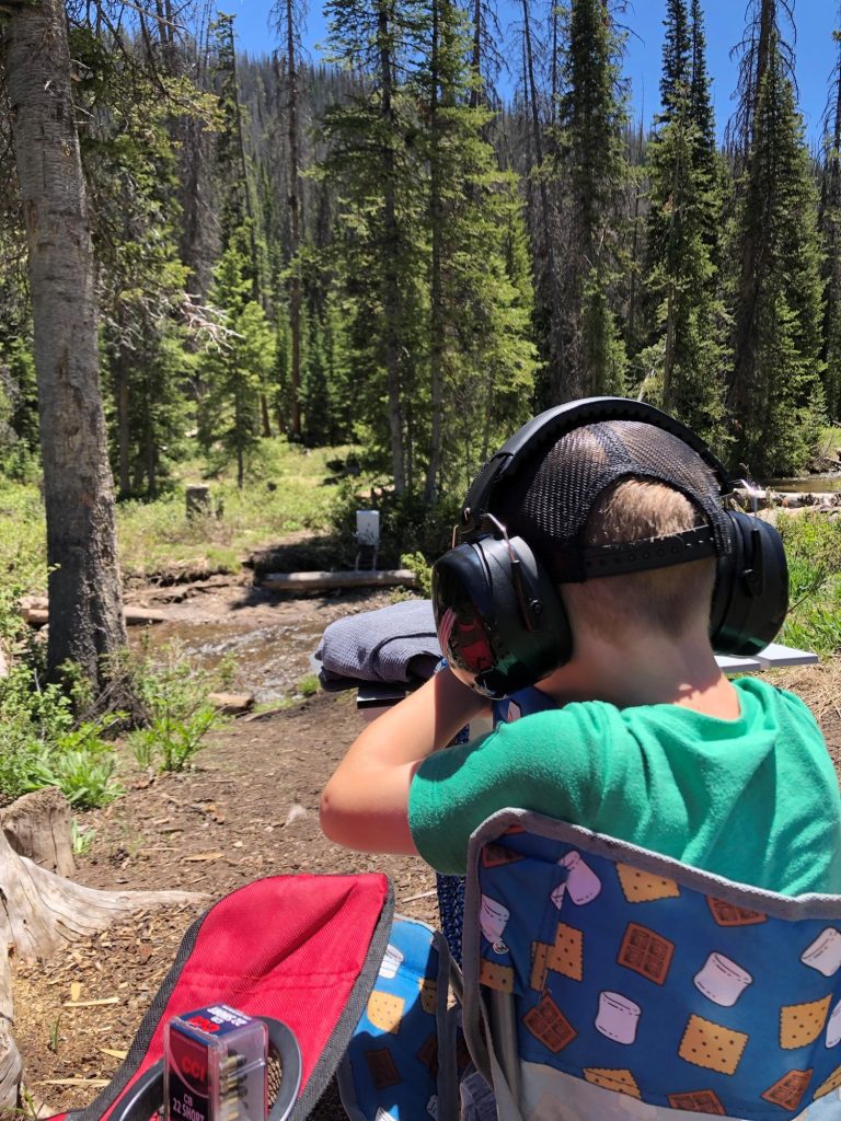 Young boy wearing ear protection and a cap aiming a rifle at a shooting target in a forest setting.